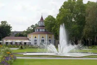 Auf dem Foto ist ein Brunnen vor einem Gebäude im Ebertpark zu sehen. Die Außenszene umfasst einen Brunnen, ein Gebäude, Pflanzen, Himmel, Bäume, Gras und Wolken.