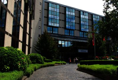 Das Bild zeigt ein Hilton-Gebaude in Mainz mit Glasfront. Es handelt sich um ein Geschaftsgebaude mit Ausenbereich, Baumen und klarem Himmel.