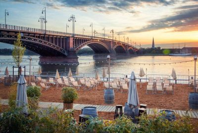 Das Bild zeigt eine Gruppe von Tischen und Stuhlen, die auf einem Dock in Mainz aufgestellt sind. Die Kulisse umfasst einen Blick auf den Fluss, eine Brucke und die Stadtlandschaft im Hintergrund. Der Himmel zeigt Anzeichen eines Sonnenaufgangs oder eines Sonn