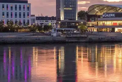 Das Bild zeigt ein Gewässer mit Gebäuden am Ufer in Ludwigshafen, darunter ein Hotel. Die Szene fängt die Außenumgebung mit Wolken am Himmel, dem See, in dem sich die Skyline spiegelt, und einer Stadtlandschaft im Hintergrund ein, die bei Sonnenaufgang oder Sonnenuntergang sichtbar ist.