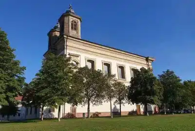 Auf dem Bild ist ein Gebäude mit Bäumen davor zu sehen, das als Ludwigshafener Kirche identifiziert wird. Die Szene umfasst Außenelemente wie Gras und den Himmel.