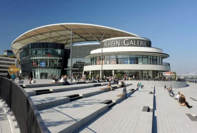 Das Bild zeigt das Gebäude der Rhein-Galerie in Ludwigshafen mit Menschen draußen. Es fängt ein großes Gebäude mit einer Stadtlandschaft im Hintergrund ein.