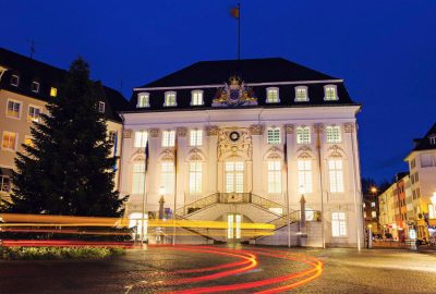 Das Bild zeigt das Alte Rathaus in Bonn. Es ist ein großes Gebäude mit einem Baum und Straßenlaternen. Das Foto wurde nachts aufgenommen und zeigt das beleuchtete Gebäude.