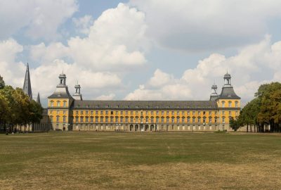 Das Bild zeigt das Kurfürstliche Schloss in Bonn, ein großes Gebäude mit einer großen Rasenfläche davor. Die Szene umfasst einen blauen Himmel mit Wolken, Gras und Bäumen. Das Gebäude ähnelt einem Palast oder einer Burg und ist in eine Stadtlandschaft eingebettet.