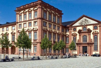 Das Bild zeigt die Mannheimer Schlosskirche in Deutschland. Es verfügt über ein großes Gebäude mit Bäumen davor. Die Architektur wirkt großartig und imposant vor einem klaren Himmel mit Wolken.