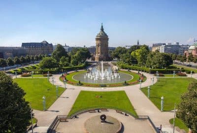 Das Bild zeigt einen großen Innenhof im Wasserturmpark in Mannheim. Es verfügt über einen Brunnen und eine Statue in der Mitte, umgeben von Gebäuden, Pflanzen und Bäumen. Die Kulisse scheint ein Stadtplatz oder ein Stadtplatz mit Landschaftselementen zu sein.