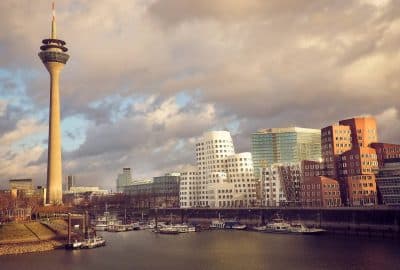 Das Foto zeigt die Skyline von Dusseldorf mit einem Fluss, einem hohen Turm und einer Brucke. Das Bild fangt die Stadtlandschaft und die durch die Stadt verlaufende Wasserstrase ein.