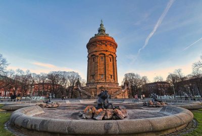 Das Bild zeigt einen Wasserturm in Mannheim, Deutschland. Vor dem Turm steht eine Statue. Die Szene umfasst einen bewölkten Himmel und einige Bäume im Hintergrund.