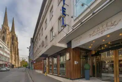 Das Bild zeigt ein Gebäude mit der Bezeichnung „HOTEL/HERZLICH“. Es befindet sich in einer städtischen Umgebung, umgeben von einer befahrenen Straße. Die Architektur spiegelt ein für Stadtstraßen typisches, gemischt genutztes Geschäftsgebäude wider.