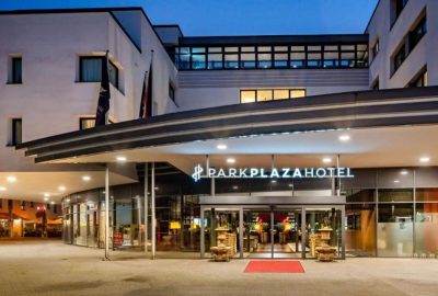 Das Bild zeigt das Hotel Park Plaza in Trier. Es zeigt ein Gebäude mit einem auffälligen roten Teppich und einer Matte, die sein elegantes Erscheinungsbild unterstreichen. Die Szenerie ist im Freien bei Nacht zu sehen und hebt die Hotelarchitektur vor dem Himmel hervor.