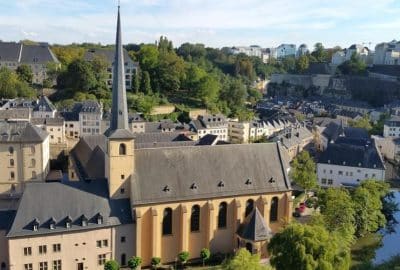 Das Bild zeigt die Janke-Kirche in Luxemburg, ein Gebäude, das sich durch seinen markanten Kirchturm auszeichnet und von Bäumen umgeben ist. Die Umgebung weist typische Elemente mittelalterlicher Architektur auf, darunter einen burgähnlichen Turm und eine Turmspitze.