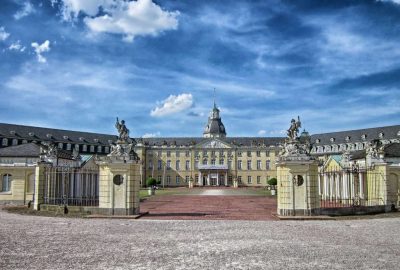 Das Bild zeigt das Karlsruher Schloss, ein großes Gebäude mit einer Statue davor. Es ist ein Steinpalast mit einer Statue im Vordergrund.