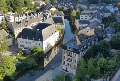 Das Bild zeigt einen ruhigen Fluss, der zwischen Gebäuden und Bäumen in einem Wohngebiet fließt. Es fängt eine Luftaufnahme der Stadtlandschaft Luxemburgs ein.