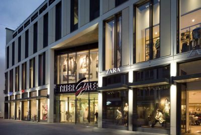 Das Bild zeigt ein Geschäftsgebäude namens Trier Galerie in Deutschland. Es präsentiert ein Schaufenster im Freien mit markanter Fassade und großen Fenstern.
