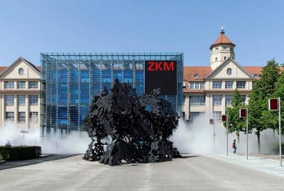 Das Bild zeigt ein großes Glasgebäude mit einem Springbrunnen davor. Das Gebäude ist das ZKM (Zentrum für Kunst und Medien). Es verfügt über eine Außenumgebung mit klarem Himmel, Bäumen und einer Straße.