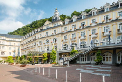 Das Bild zeigt das Kurhaus Bad Ems, ein großes Gebäude, das sich durch seine markante Fassade und architektonische Gestaltung auszeichnet. Es verfügt über einen Gehweg, der zum Eingang führt, sowie über Treppen, die seine Pracht unterstreichen.