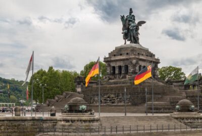 Das Bild zeigt die Statue eines Mannes auf einem Pferd auf einer Steinkonstruktion, die Teil der Gedenkstätte Deutsche Einheit in Koblenz ist. Die Szene spielt im Freien vor einem klaren Himmel mit Wolken und zeigt Flaggen in der Nähe.