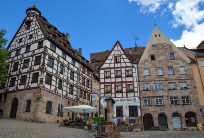 Die Szene spielt im Freien bei leicht bewölktem Himmel. Sie zeigt eine architektonische Kulisse, die wahrscheinlich zu Nürnberg gehört, und präsentiert die historischen Regierungsgebäude der Stadt sowie die Bäume in der Umgebung.