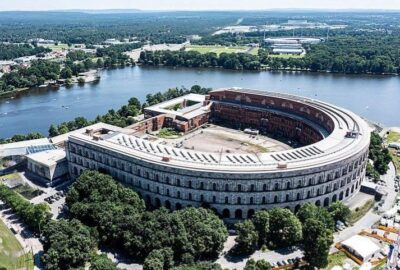 Das Bild zeigt die Nürnberger Aerial Kongresshalle, ein kreisförmiges Gebäude, umgeben von Bäumen. Es ist aus der Vogelperspektive aufgenommen und zeigt das Design des Bauwerks in einer städtischen Umgebung.