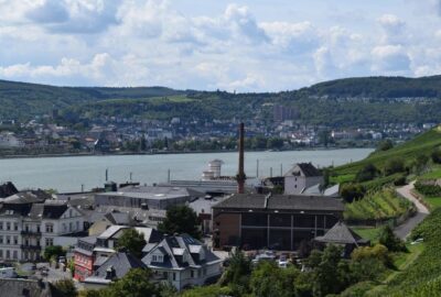 Das Bild zeigt eine charmante Stadt am Fluss, Rüdesheim am Rhein in Deutschland. Es zeigt eine malerische Landschaft mit Gebäuden am Ufer, umgeben von üppigem Grün und Bäumen.