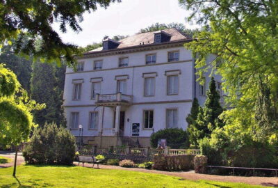 Das Bild zeigt ein großes weißes Gebäude, wahrscheinlich ein historisches Herrenhaus oder Gutshaus, umgeben von Bäumen und gepflegtem Rasen.