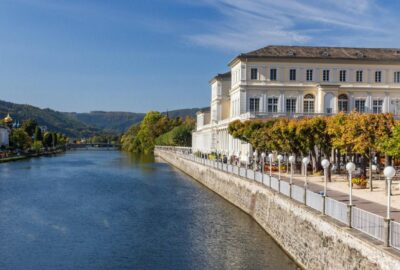 Das Bild zeigt das Kurtheater in Bad Ems, direkt an einem Gewässer gelegen. Das Gebäude ist von natürlichen Elementen wie Bäumen und Bergen umgeben.