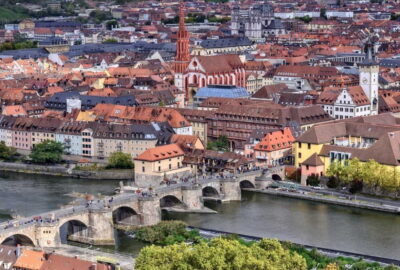 Das Bild zeigt die Alte Mainbrücke in Deutschland, die sich über einen Fluss erstreckt. Im Hintergrund sind verschiedene Gebäude und eine Kirche zu sehen.