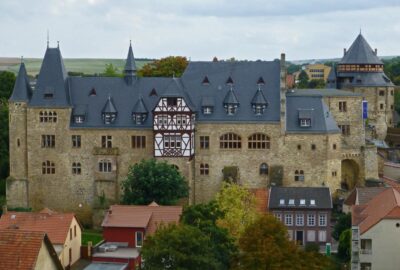 Das Bauwerk zeichnet sich durch Türmchen und ein markantes Dach aus, das typisch für die Burgarchitektur ist. Es steht vor einer Kulisse aus Bäumen und Wolken am Himmel und befindet sich in einer Stadtumgebung.