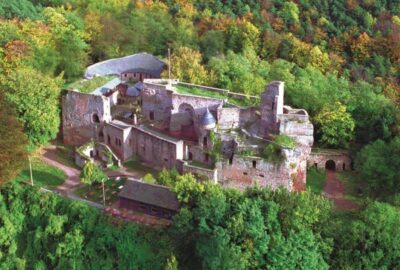 Das Bild zeigt eine alte Burg in Landstuhl, Deutschland, umgeben von üppigen Bäumen und Grün. Die Burg liegt auf einem Hügel und unterstreicht ihre majestätische Präsenz in der ländlichen Landschaft.