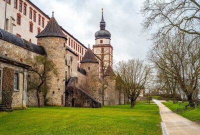 Das Bild zeigt die Festung Marienberg, eine historische Festung in Würzburg. Das Gebäude besticht durch mittelalterliche Architektur mit Türmen und Mauerwerk und ist von üppigem Rasen und Bäumen umgeben.