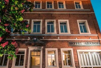 Das Bild zeigt das Hotel Arador, ein Gebäude, das sich durch seine vielen Fenster auszeichnet. Es zeigt eine Fassade, die für den Außenbereich konzipiert ist, mit sichtbaren Elementen wie Türen und Blumenarrangements.