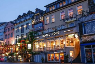 Das Bild zeigt eine Reihe von Gebäuden, insbesondere das Hotel Noss, das nachts beleuchtet ist. Es befindet sich in einem gemischt genutzten Gebiet mit sichtbaren Geschäften und Fenstern, was eine lebendige Atmosphäre in der Stadt schafft.