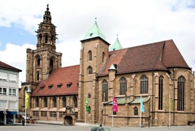Das Bild zeigt die Kiliansgemeinde, ein großes Gebäude mit einem markanten Turm. Die Szenerie ist im Freien, unter einem klaren Himmel mit Wolken, in der Stadt Heilbronn, Deutschland, zu sehen.