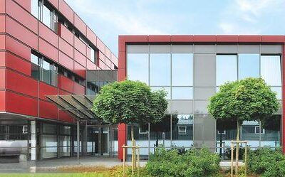 Das Bild zeigt ein Gebäude in Alzey, Deutschland, vor dem Bäume stehen. Die sichtbare Fassade besteht aus Fenstern und einer Struktur, die zu Immobilien- oder Verwaltungsfunktionen zu gehören scheint.