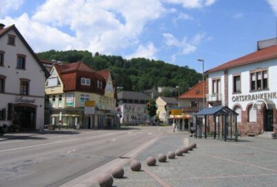 Das Bild zeigt eine Straßenszene in Landstuhl, Deutschland, mit verschiedenen Gebäuden entlang der Straße. Die Umgebung ist im Freien mit klarem Himmel und Wolken.