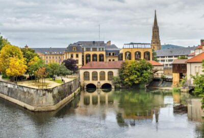 Das Bild zeigt eine malerische Ansicht von Metz in Frankreich mit einem Gewässer, das von Gebäuden und Bäumen umgeben ist.