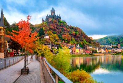 Das Bild zeigt einen malerischen Pfad, der zu einem ruhigen See führt, mit einem malerischen Schloss auf einem Hügel im Hintergrund.