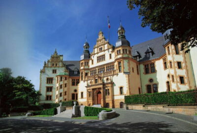 Das Bild zeigt einen großen Palast, der sich durch sein beeindruckendes Dach und seine mittelalterliche Architektur auszeichnet. Das Bauwerk steht vor einer Außenkulisse mit klarem Himmel und einigen Wolken.