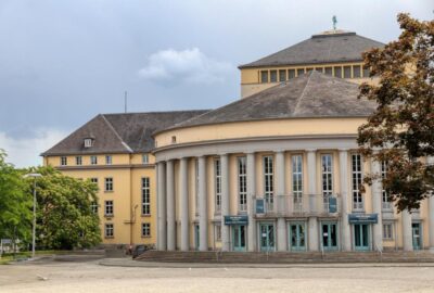 Das Bild zeigt das Saarländische Staatstheater, ein markantes Gebäude, das durch seine Säulen und sein rundes Dach charakterisiert ist.