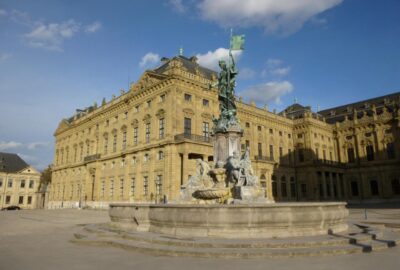 Das Bild zeigt eine Statue, die prominent vor einem bedeutenden Gebäude in Würzburg steht. Das Bauwerk präsentiert klassische Architektur und dient als Wahrzeichen.